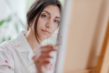 Foto de Joven artista femenina confiada pintando sobre lienzo, sosteniendo a mano el pincel en primer plano - Imagen libre de derechos