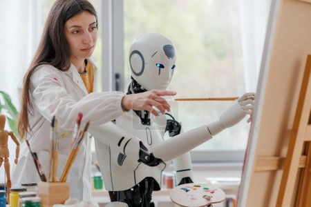 Foto de Artista mujer enseñando pintura a un robot humanoide AI, ella está apuntando a lienzo y dando consejos - Imagen libre de derechos