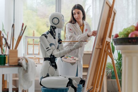 Foto de Artista mujer enseñando arte y pintura a un robot humanoide AI, ella está apuntando a la lona y dando consejos - Imagen libre de derechos