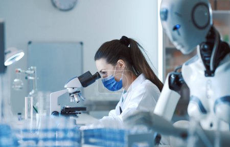 Photo for Scientist and AI robot working together in the lab: the woman is looking at the samples under the microscope and the robot is assisting her - Royalty Free Image