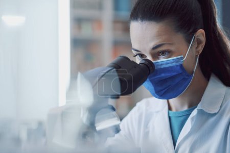 Foto de Científica que trabaja en el laboratorio, lleva una máscara quirúrgica y un microscopio. - Imagen libre de derechos