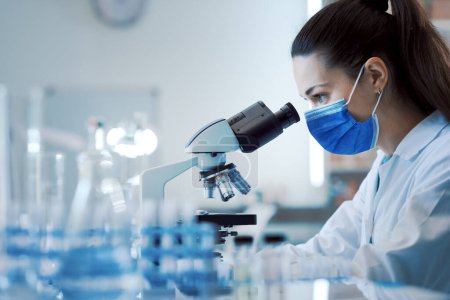Female scientist working in the lab, she is wearing a surgical mask and using a microscope