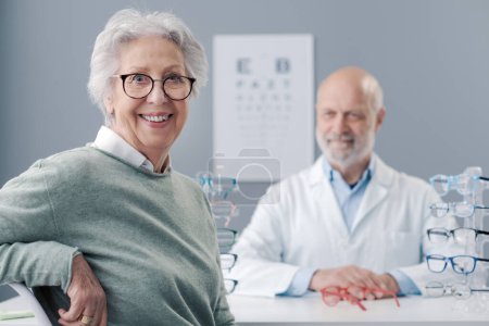 Téléchargez les photos : Heureuse femme âgée qui choisit ses nouvelles lunettes de vue, la spécialiste des soins oculaires l'aide - en image libre de droit