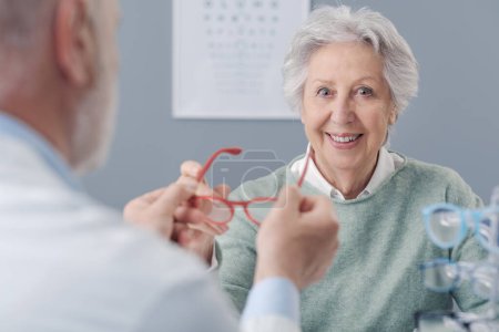 Foto de Mujer anciana sonriente probando nuevas gafas graduadas, el oculista la está ayudando - Imagen libre de derechos