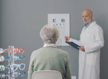 Téléchargez les photos : Femme âgée ayant un examen de la vue avec un ophtalmologiste professionnel, elle est assise et regarde le tableau des yeux - en image libre de droit