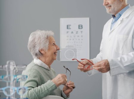 Foto de Feliz mujer mayor que se somete a un examen ocular, el médico le está dando un nuevo par de gafas graduadas - Imagen libre de derechos