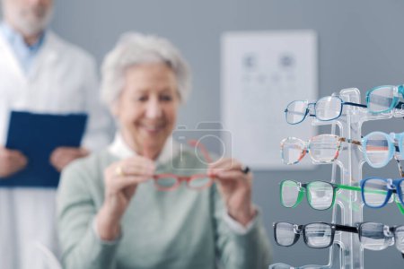 Téléchargez les photos : Femme âgée essayant de nouvelles lunettes, l'oculiste se tient près d'elle - en image libre de droit
