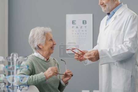 Téléchargez les photos : Heureuse femme âgée ayant un examen de la vue, le médecin lui donne une nouvelle paire de lunettes de vue - en image libre de droit