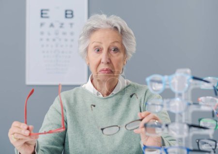 Téléchargez les photos : Femme âgée choisissant des lunettes à la boutique de lunettes, elle est confuse et coûteuse - en image libre de droit