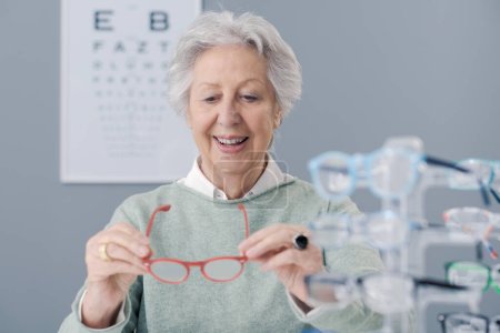 Photo for Senior woman choosing glasses at the eyewear shop, she is holding eyeglasses and smiling - Royalty Free Image