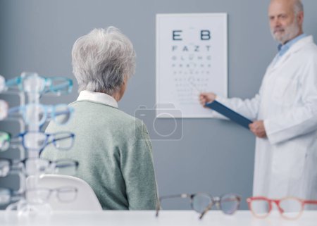 Téléchargez les photos : Femme âgée ayant un examen de la vue avec une oculiste professionnelle, elle est assise et regarde le tableau des yeux - en image libre de droit
