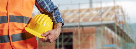 Photo for Building contractor holding a safety helmet and construction site in the background - Royalty Free Image
