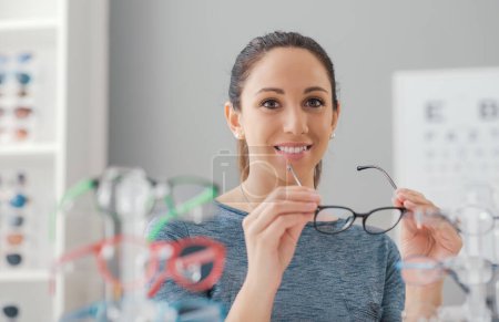 Téléchargez les photos : Jeune femme souriante choisissant une nouvelle paire de lunettes de vue au magasin de lunettes - en image libre de droit