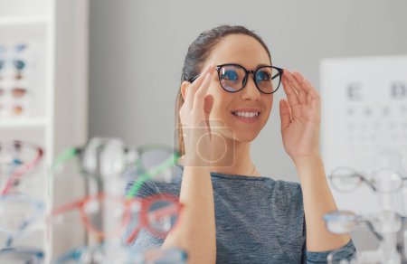 Téléchargez les photos : Jeune femme souriante choisissant une nouvelle paire de lunettes de vue au magasin de lunettes - en image libre de droit