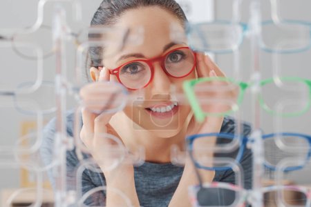 Foto de Hermosa mujer feliz en la tienda de óptica, ella está mirando la colección de gafas en la pantalla y elegir un nuevo par de gafas graduadas - Imagen libre de derechos