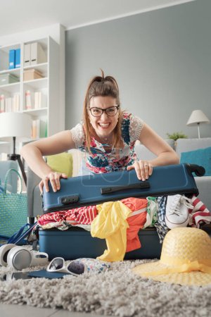 Téléchargez les photos : Stressée jeune femme préparant ses bagages pour un voyage, elle ferme sa valise trolley surchargée - en image libre de droit
