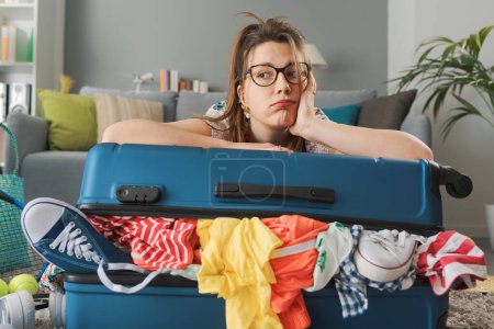 Photo for Tired sad woman packing for a long journey, she is leaning on her overfilled trolley bag: travel, vacations and tourism concept - Royalty Free Image