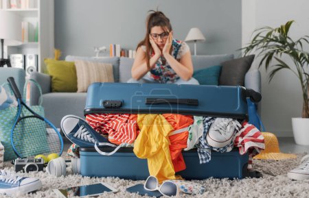Photo for Stressed woman sitting on the couch at home and big overfilled trolley bag, she is packing for a vacation - Royalty Free Image