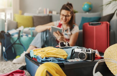 Foto de Feliz joven mujer empacando sus maletas para un viaje, se está conectando en línea con su tableta y sonriendo: viaje y concepto de vacaciones - Imagen libre de derechos