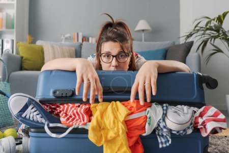 Photo for Tired sad woman packing for a long journey, she is leaning on her overfilled trolley bag: travel, vacations and tourism concept - Royalty Free Image