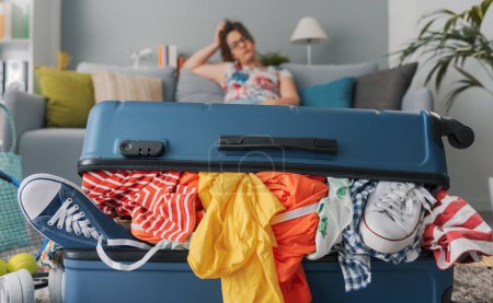 Stressed woman sitting on the couch at home and big full trolley bag, she is packing for a vacation