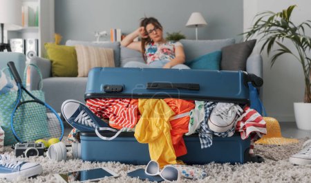 Foto de Mujer estresada sentada en el sofá en casa y gran bolsa de carro lleno, ella está empacando para unas vacaciones - Imagen libre de derechos