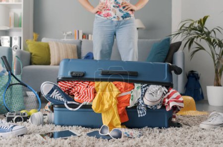 Foto de Mujer decepcionada empacando para un viaje: ella está de pie delante de su maleta llena de ropa, viajes y concepto de vacaciones - Imagen libre de derechos