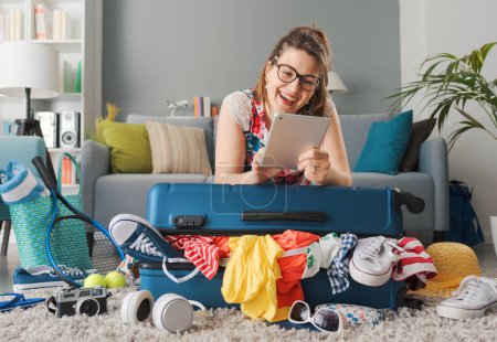 Photo for Smiling woman leaning on her trolley bag filled with clothes and connecting online with her tablet, she is preparing her luggage for a trip: travel and vacations concept - Royalty Free Image