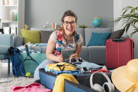 Photo for Happy young woman packing her suitcases for a trip, she is connecting online with her tablet and smiling: travel and vacations concept - Royalty Free Image