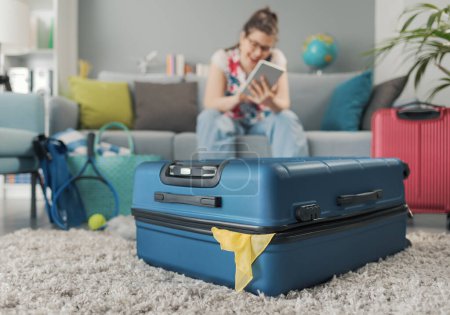 Foto de Joven viajera sentada en el sofá y conectada con su tablet, empacó su equipaje y está lista para irse. - Imagen libre de derechos