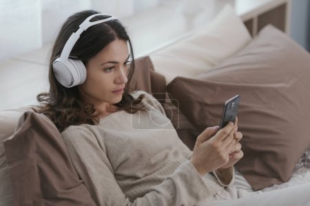 Photo for Young woman relaxing on the bed, she is wearing headphones and watching videos on her smartphone - Royalty Free Image