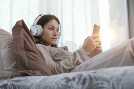 Foto de Young woman relaxing on the bed, she is wearing headphones and watching videos on her smartphone - Imagen libre de derechos