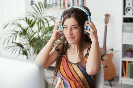 Photo for Young woman sitting at desk at home, she is watching the computer screen and wearing headphones - Royalty Free Image