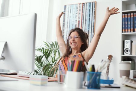 Photo for Cheerful successful woman celebrating with arms raised, she is sitting at desk at home and connecting with her computer - Royalty Free Image