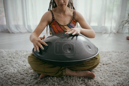 Téléchargez les photos : Femme créative assise sur le sol à la maison et jouant un tambour suspendu, concept de musique et d'arts - en image libre de droit