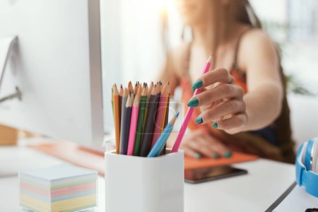 Photo for Young designer sitting at desk and working, she is taking a colored pencil, creativity and imagination concept - Royalty Free Image