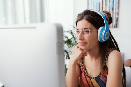 Téléchargez les photos : Jeune femme assise au bureau à la maison, elle regarde l'écran de l'ordinateur et porte des écouteurs - en image libre de droit