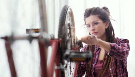 Foto de Mujer confiada arreglando su propia bicicleta en casa, está usando una llave inglesa y chequeando ruedas - Imagen libre de derechos