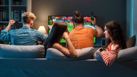 Téléchargez les photos : Les filles ennuyées parlent et bavardent ensemble pendant que leurs copains regardent le football à la télévision - en image libre de droit