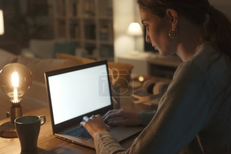 Téléchargez les photos : Femme assise au bureau à la maison et travaillant avec son ordinateur portable la nuit, elle tape sur le clavier, maquette d'écran - en image libre de droit