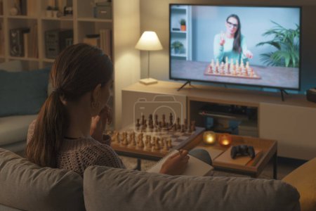 Téléchargez les photos : Femme assise sur le canapé et apprenant à jouer aux échecs, elle regarde une émission de télévision - en image libre de droit