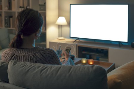 Téléchargez les photos : Femme relaxante sur le canapé à la maison et regardant la télévision, elle tient la télécommande, maquette d'écran - en image libre de droit