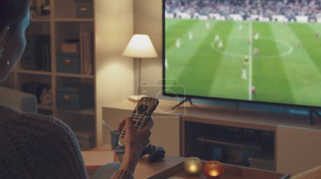 Téléchargez les photos : Femme reaxing sur le canapé et regarder un match de football sur sa télévision intelligente, elle tient la télécommande - en image libre de droit