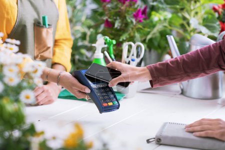 Téléchargez les photos : Client payant dans une boutique de fleurs en utilisant son smartphone, concept de paiements électroniques - en image libre de droit
