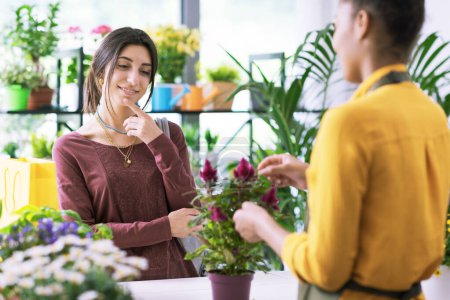 Téléchargez les photos : Jeune femme achetant une plante à fleurs à la boutique de fleurs, elle regarde la plante et pense - en image libre de droit