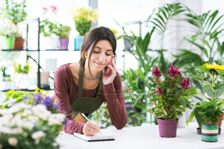 Photo for Florist working in a flower shop, she is taking orders over the phone - Royalty Free Image