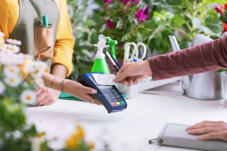 Photo for Customer at the florist shop, she is paying with her credit card, the shop owner is holding the POS terminal, hands close up - Royalty Free Image