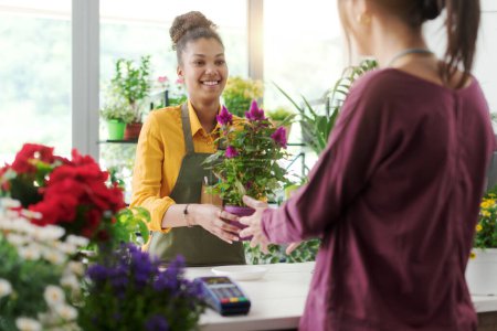 Foto de Mujer joven comprando una planta con flores en la floristería, la floristería está sosteniendo una hermosa planta - Imagen libre de derechos