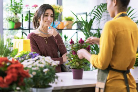 Photo for Young woman buying a flowering plant at the flower shop, she is looking at the plant and thinking - Royalty Free Image