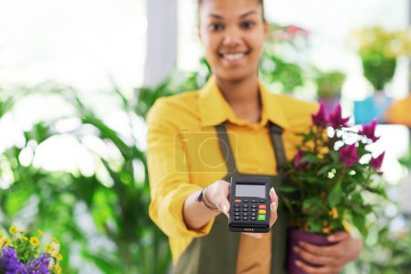 Téléchargez les photos : Jeune fleuriste souriant tenant un terminal POS et regardant la caméra, concept de paiements électroniques - en image libre de droit